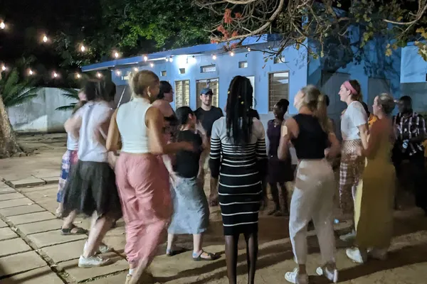 A group of people dancing together outside at night, under string lights, with a blue building in the background.