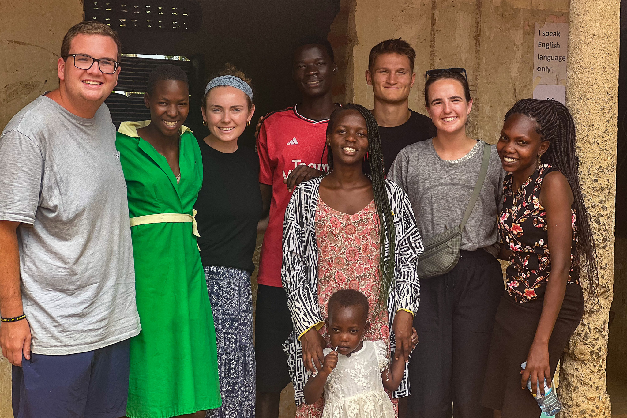 A diverse group of people, including a child, smiling and standing together in front of a rustic building.
