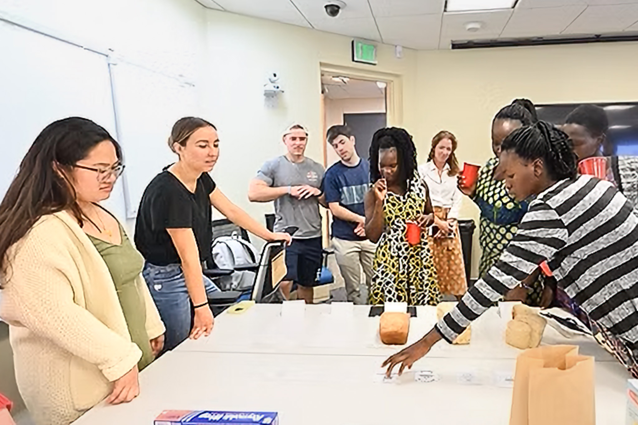 A group of people stands around a table with various loaves of bread, in a room with a whiteboard and overhead lighting.