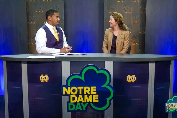 Two people sit at a news desk with Notre Dame Day signs and logos, engaged in conversation.