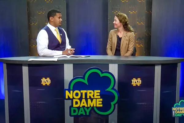 Two people sit at a news desk with Notre Dame Day signs and logos, engaged in conversation.