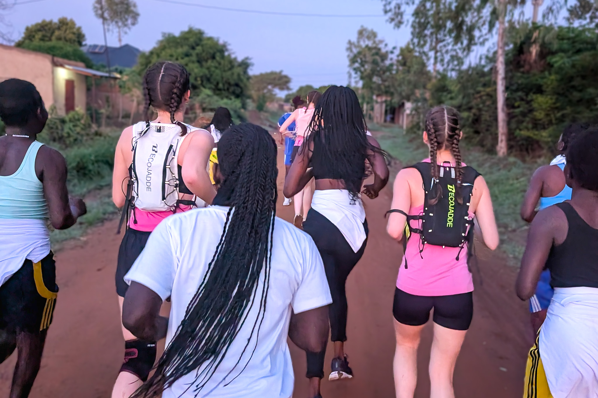 A group of people with braided hair run on a dirt path through a rural area, surrounded by trees and buildings.