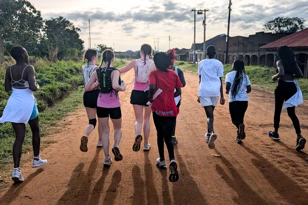 A group of people running together on a dirt road in a rural area, under a cloudy sky.