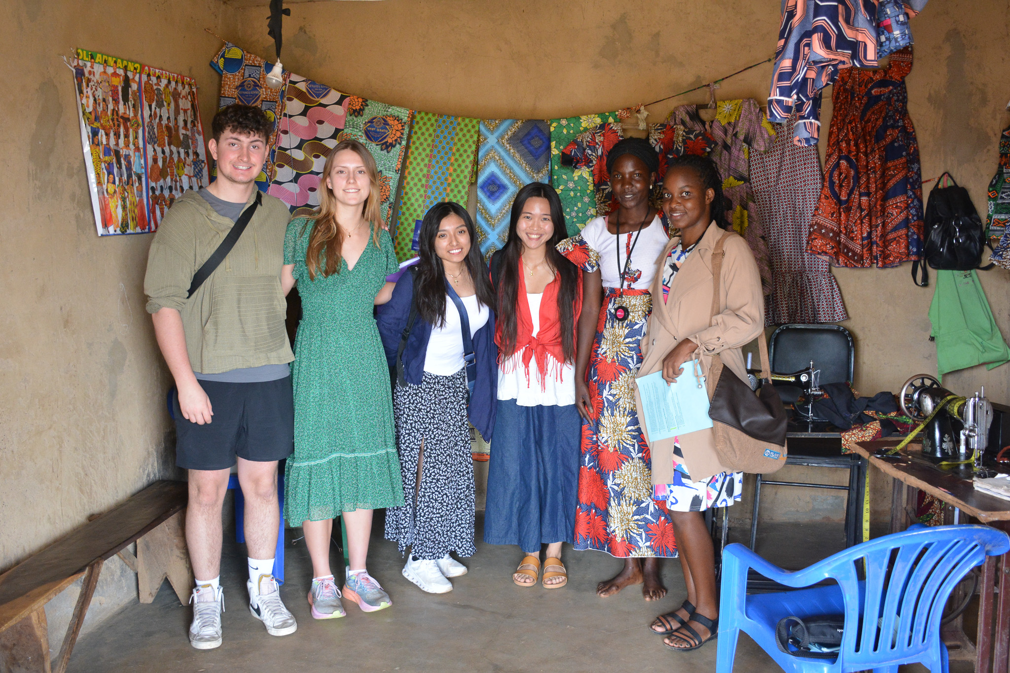 A group of six people smiling inside a colorful fabric shop, with sewing equipment and vibrant textiles in the background.
