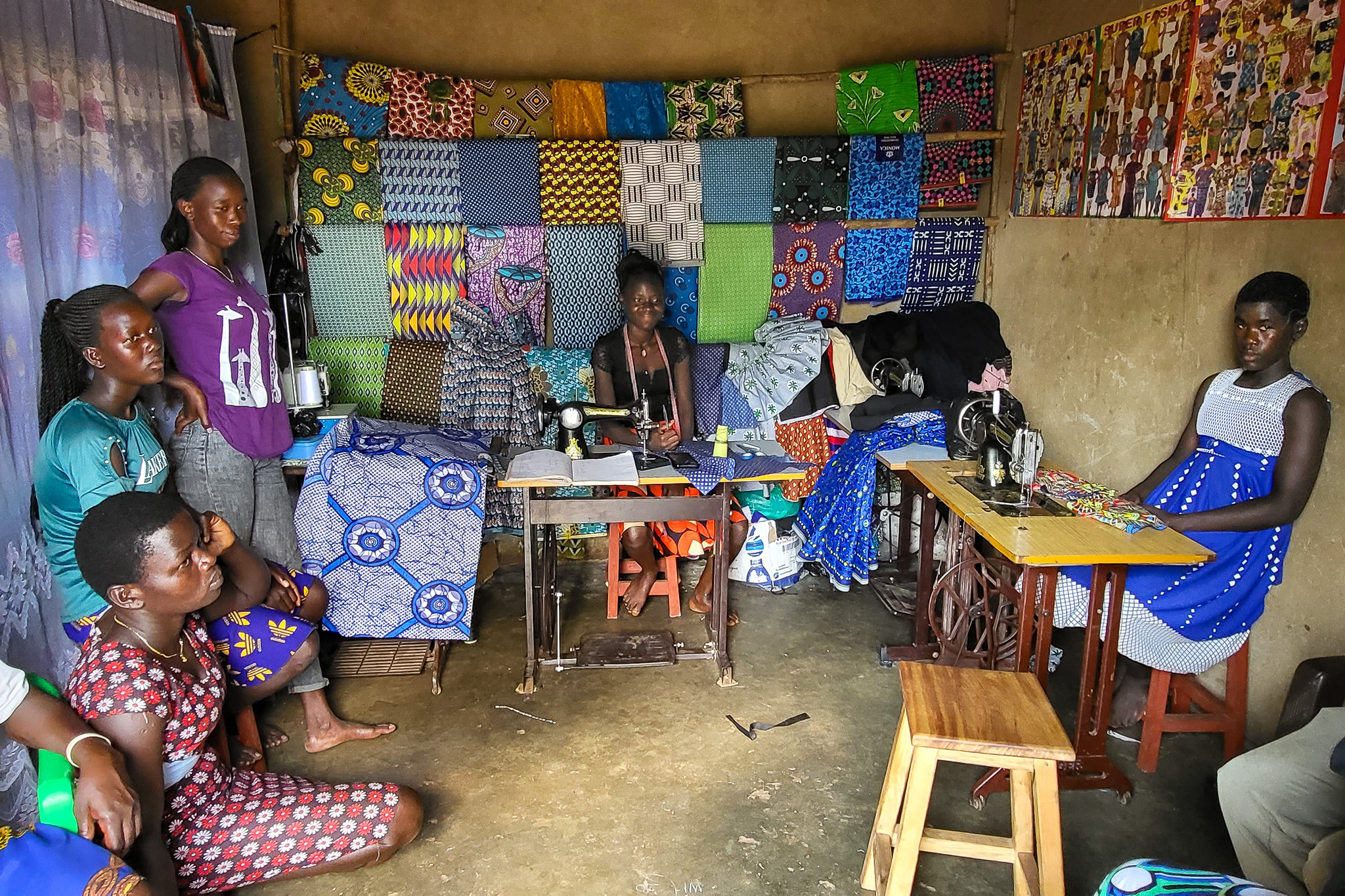A group of women in a colorful fabric shop, some sewing, others sitting, surrounded by vibrant textiles on the walls.
