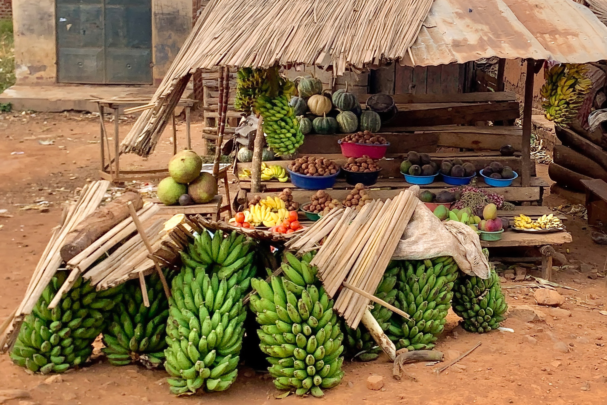 Outdoor fruit stand with bunches of bananas, melons, and various fruits in baskets under a makeshift wooden shelter.