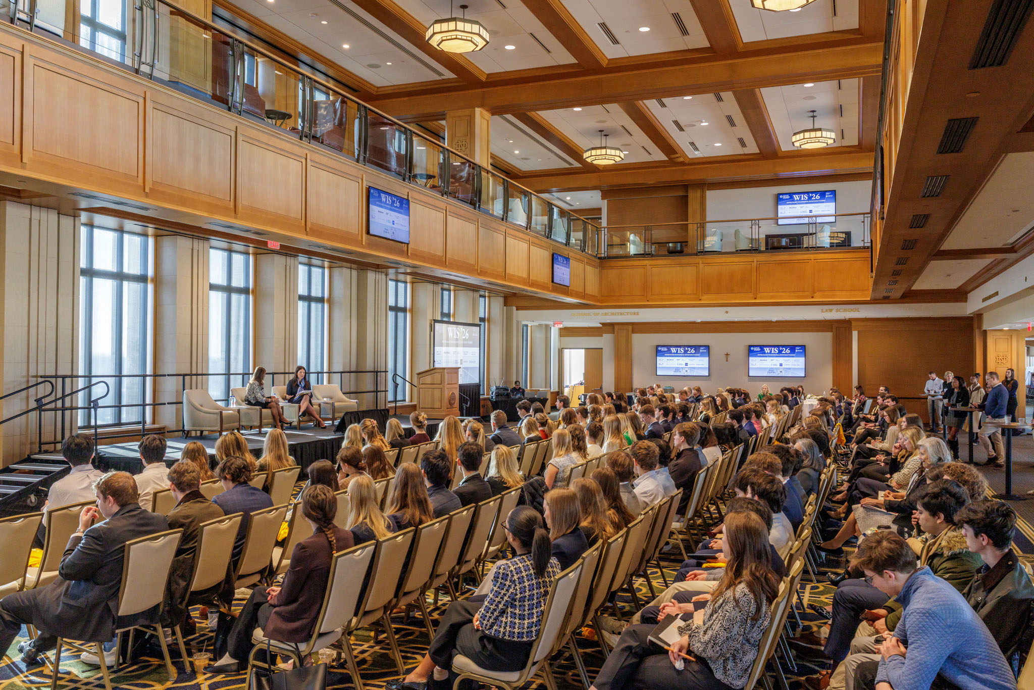 WIS '26 Keynote Conference room with attendees seated, facing a stage with two speakers. Large windows and screens display "WIS '26" event branding.