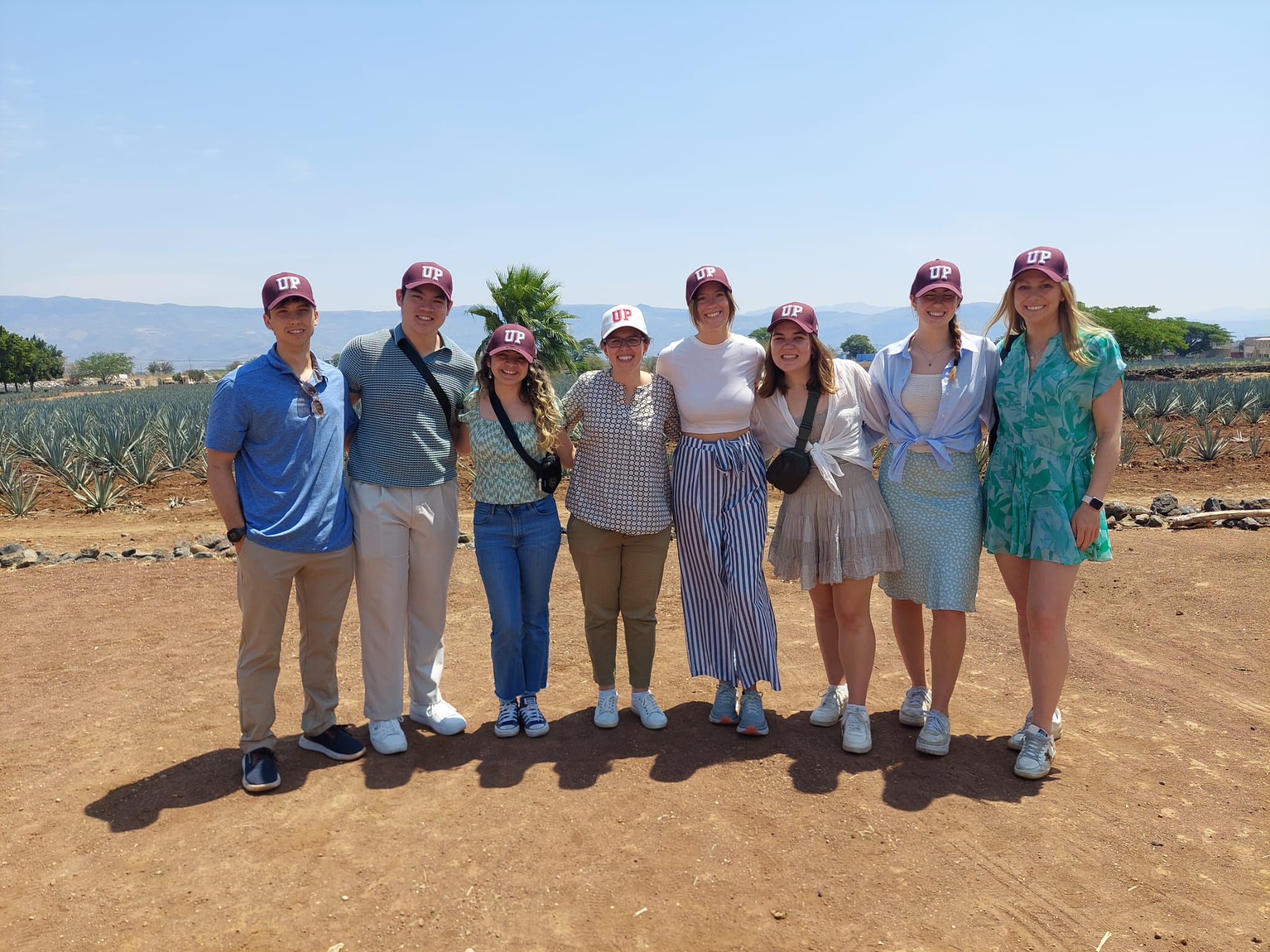 A group of eight people wearing matching maroon "UP" hats, standing in a line outdoors on a sunny day with agave plants in the background.