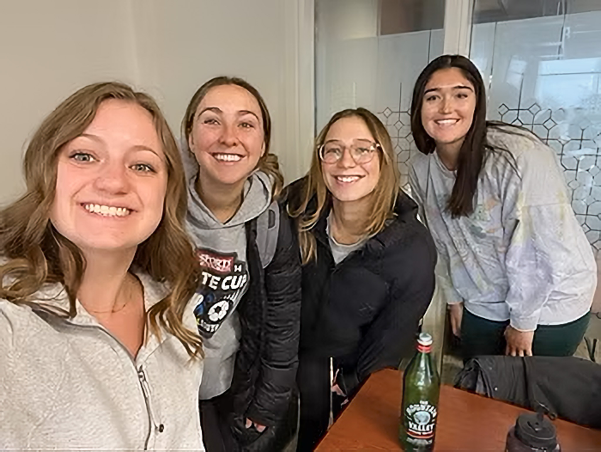 Four smiling women posing indoors, standing around a table, against a glass wall with decorative patterns.