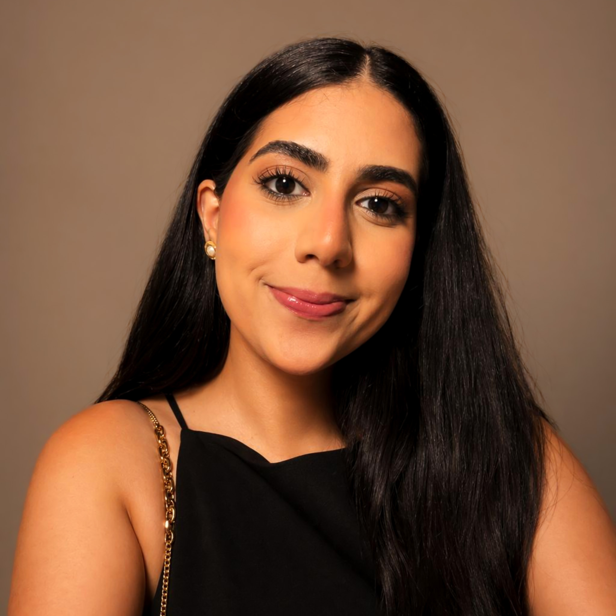 Smiling woman with long black hair, wearing a black top and gold earrings, against a plain background.