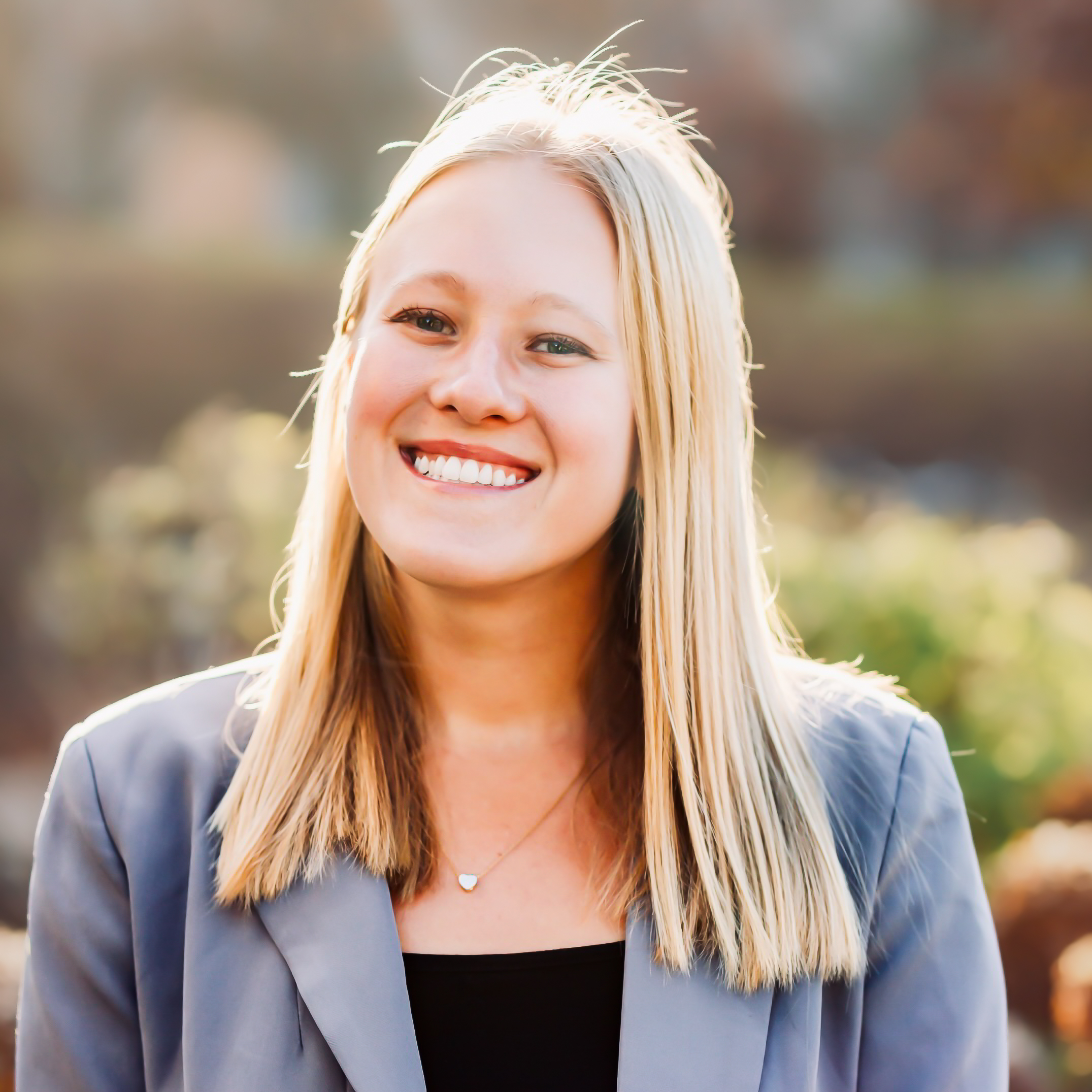 Smiling person with long blonde hair in a gray blazer, standing outdoors with a blurred natural background.