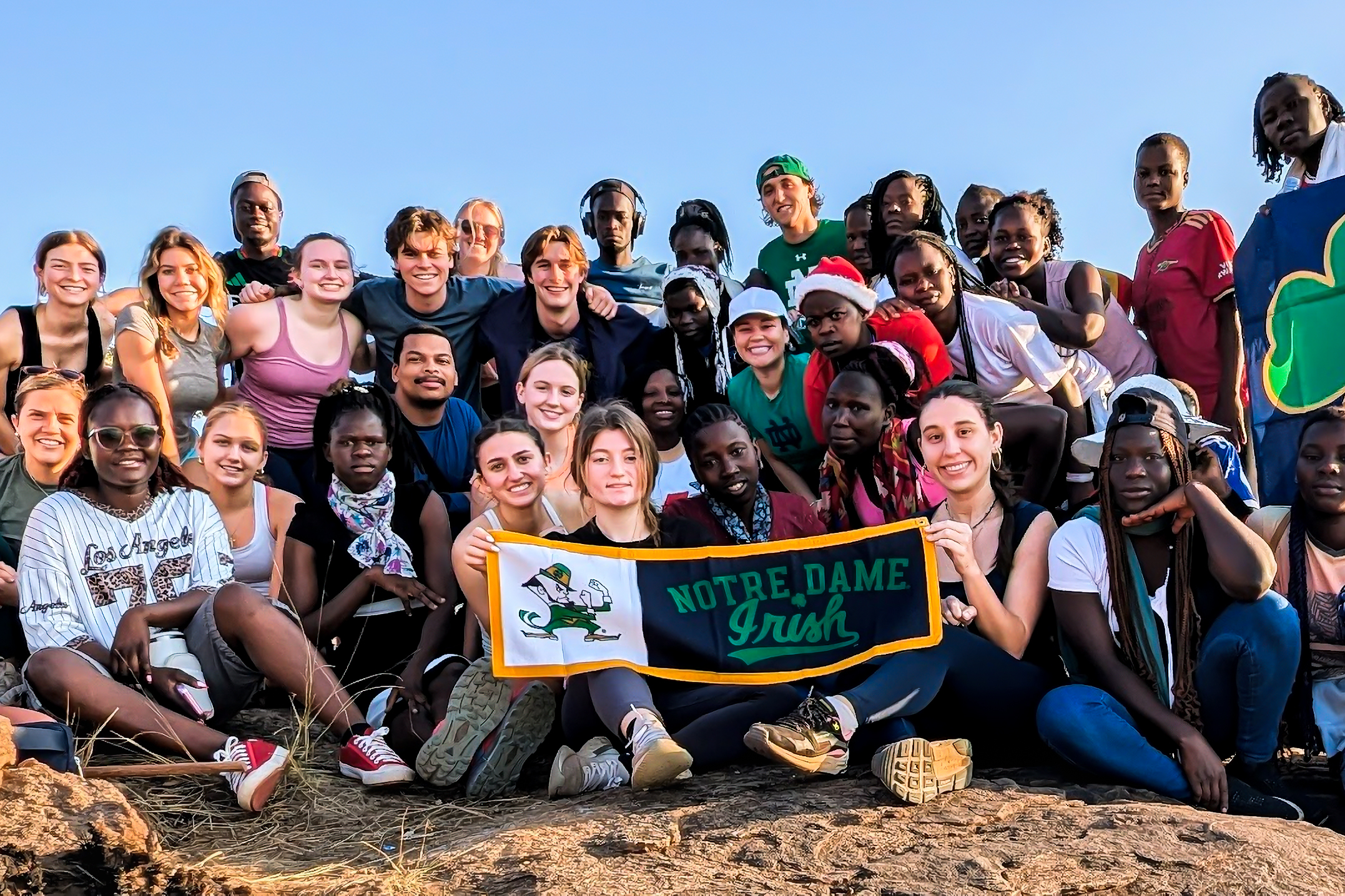 A diverse group of smiling people poses outdoors, holding a "Notre Dame Irish" banner under a clear blue sky.