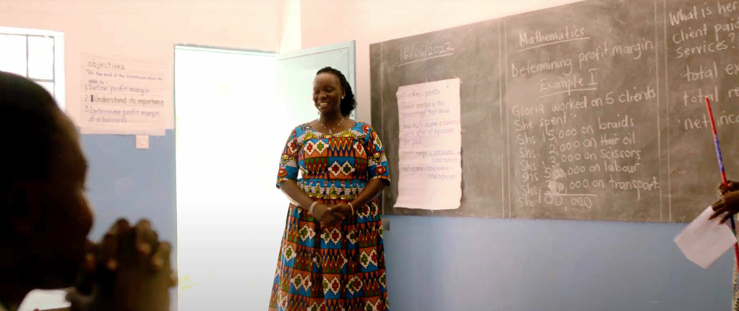 A woman in a colorful dress stands in front of a classroom, smiling, with a chalkboard displaying math problems behind her.