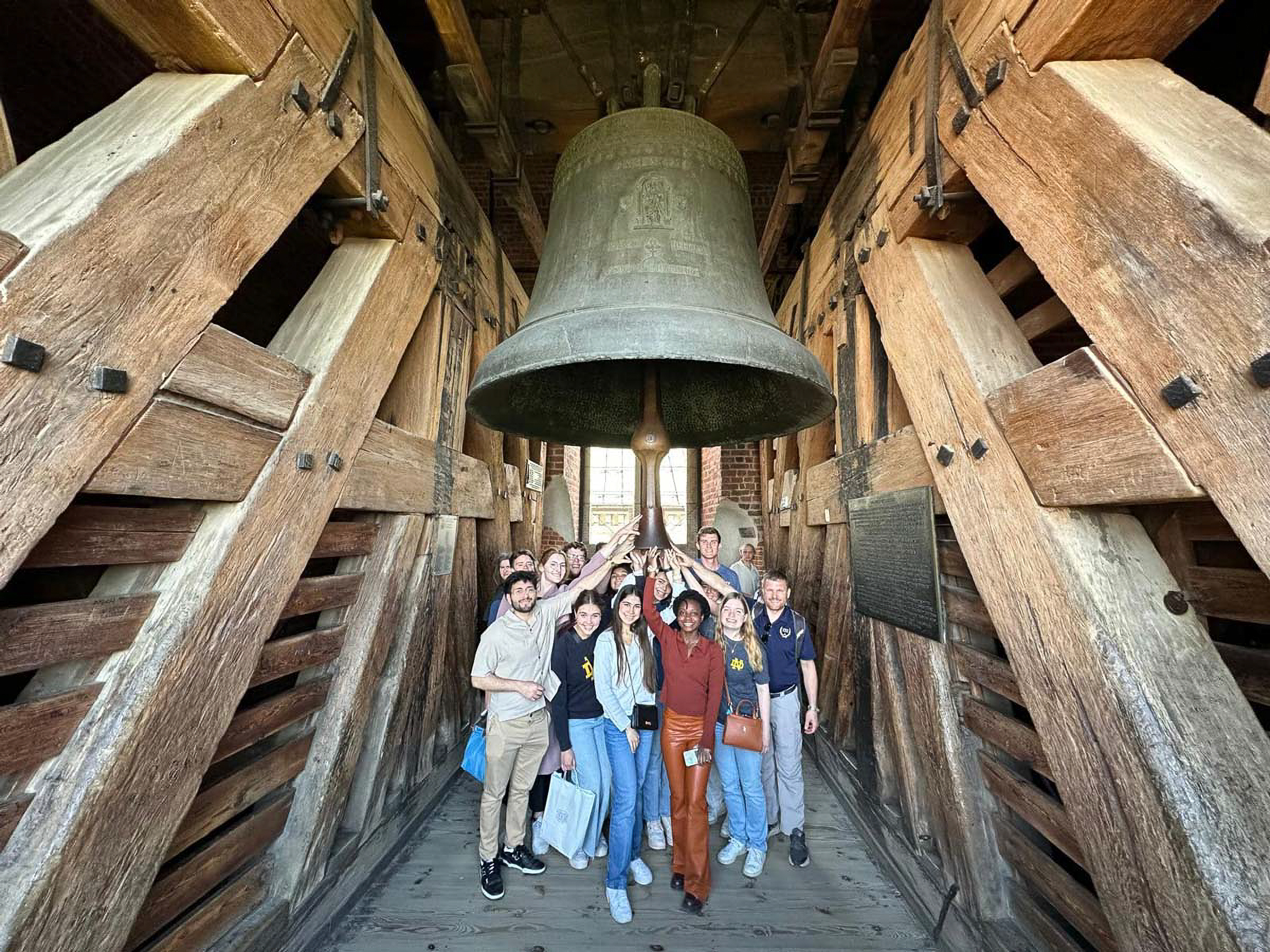 A group of people stands around a large historic bell inside a wooden structure, posing cheerfully and pointing towards the bell.