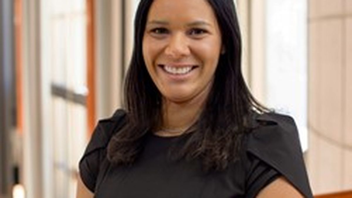Smiling Meghan Huff with long dark hair in a black dress stands indoors with a modern, bright background.