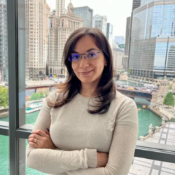 A person with long hair and glasses stands by a window overlooking a cityscape with tall buildings and a river.