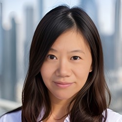 A person with long dark hair and a white shirt stands in front of a blurred cityscape background, smiling slightly at the camera.