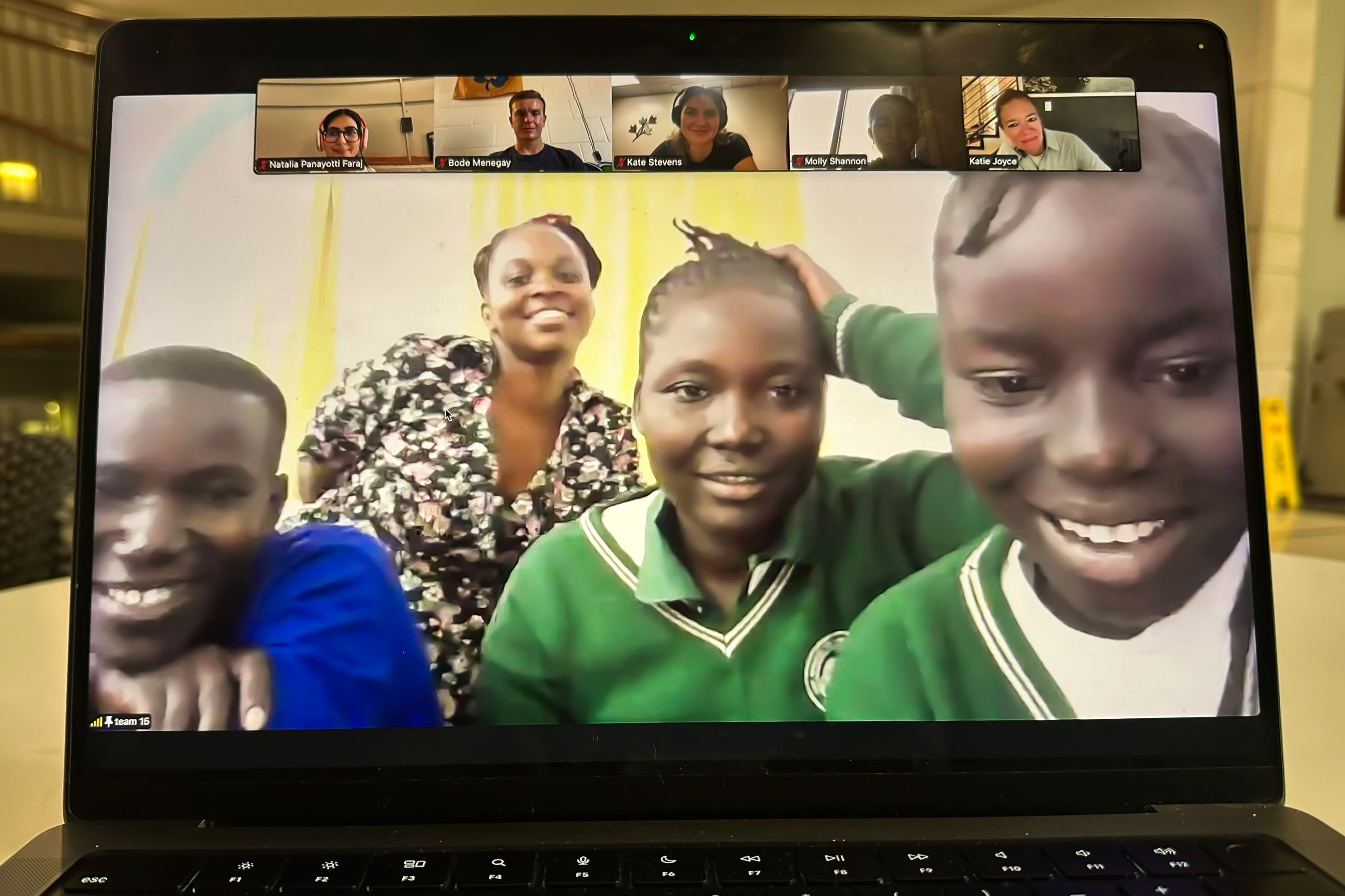 A laptop screen displays a video call with smiling participants, including four individuals in green uniforms and several others in small windows above.