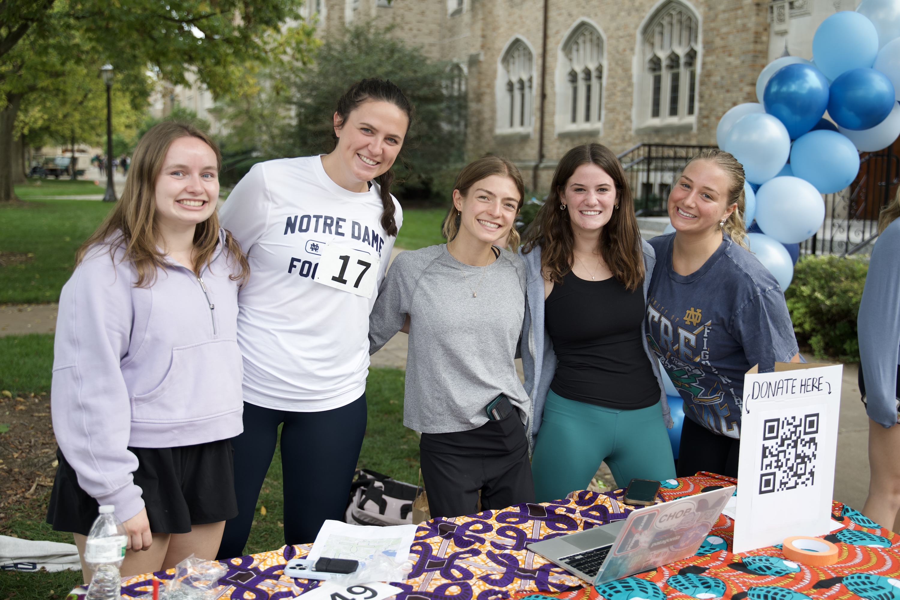 Five young women smile at an outdoor donation booth with balloons, a QR code sign, and a laptop.