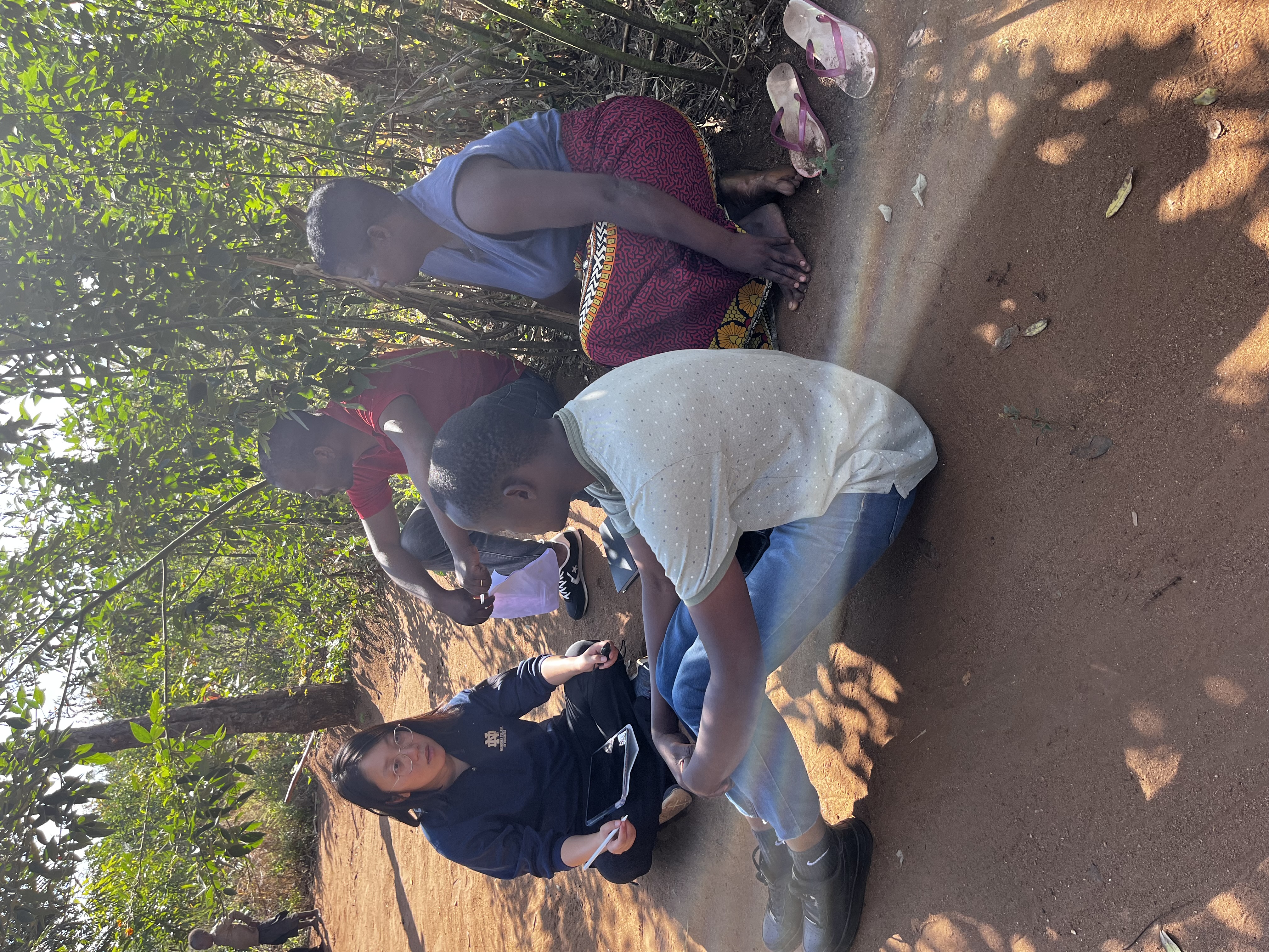 A group of four people sitting and talking on a dirt pathway, surrounded by greenery. One person is holding a tablet.