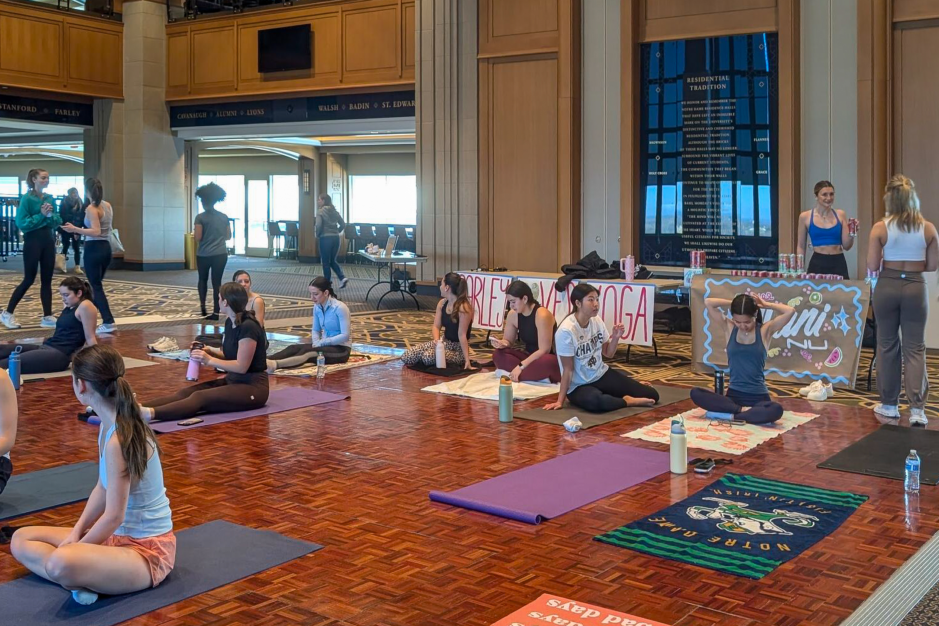 People practicing yoga on mats indoors, surrounded by banners and refreshments. The room is spacious with a decorative floor and walls.