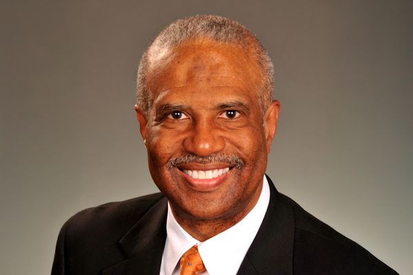 Smiling man in a suit and orange tie against a gray background.