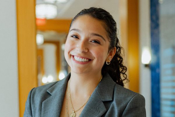 Smiling woman with curly hair in a gray blazer, wearing gold necklaces, stands in a well-lit room with wooden accents.