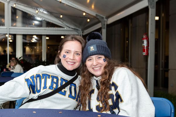 Two people wearing Notre Dame sweatshirts, smiling and sitting at a table in a covered outdoor area. One wears a beanie with a pom-pom.