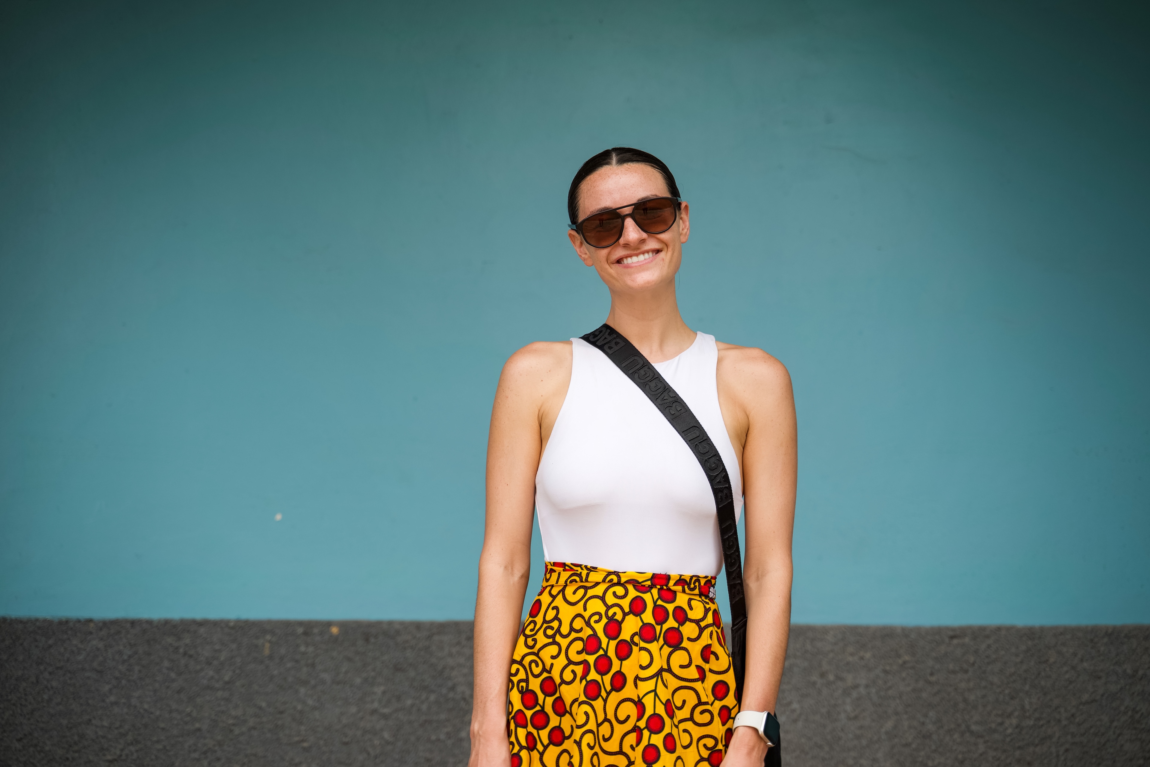 Smiling person in sunglasses, wearing a white top and colorful skirt, stands against a blue and gray background.