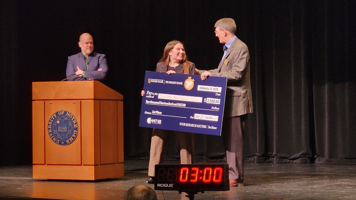 Three people on stage at Notre Dame, one holding a large check for $250. Podium and timer in the foreground, black curtain in the background.
