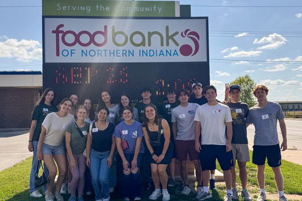 A group of people posing in front of a "Food Bank of Northern Indiana" sign on a sunny day. The sign reads "Serving the Community."