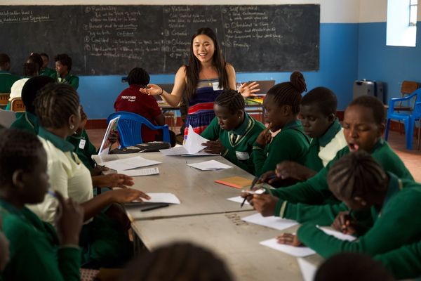 A teacher engages with students in a classroom. Students in green uniforms are seated at tables, writing and discussing. A chalkboard is in the background.