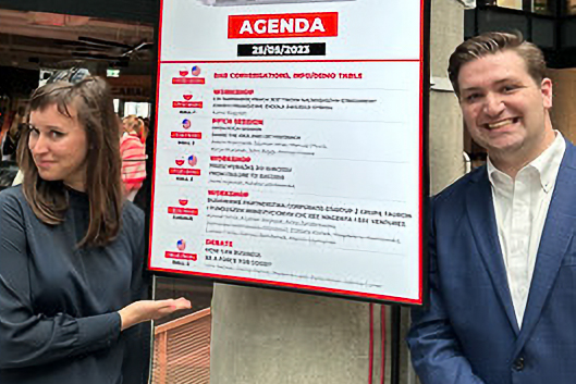 Two people stand beside an agenda poster. The woman gestures towards the poster.