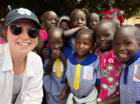 A smiling person in sunglasses and a cap poses with a group of cheerful children in blue uniforms, outdoors under trees.