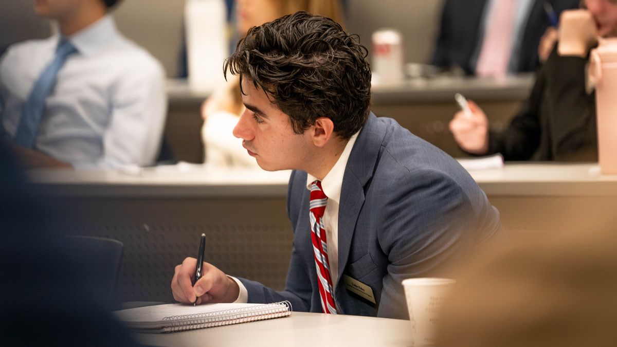 A young man in a suit and striped tie takes notes attentively in a lecture room, surrounded by other focused individuals.