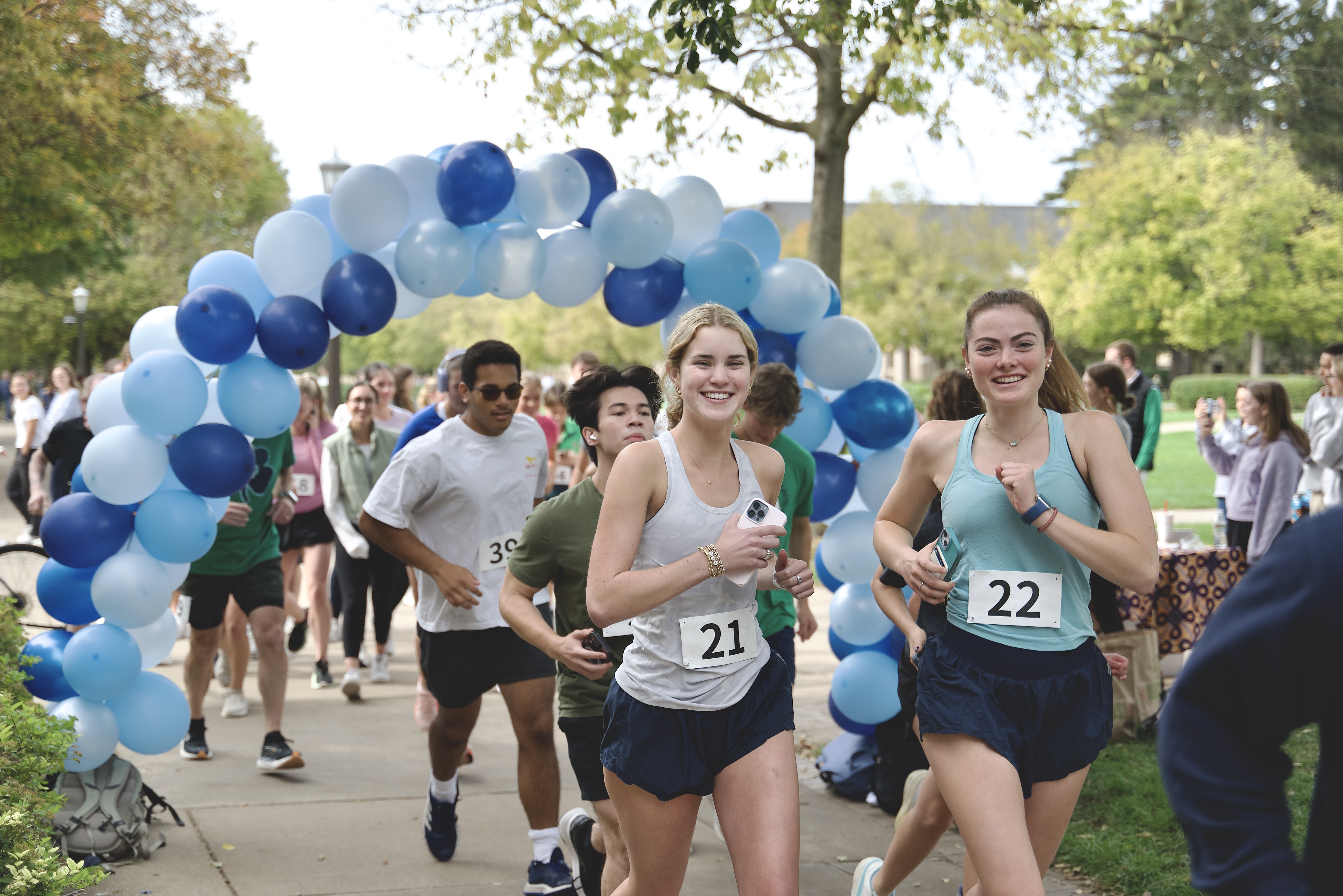 Runners with race numbers 21 and 22 smiling as they pass under a blue and white balloon arch during an outdoor event.