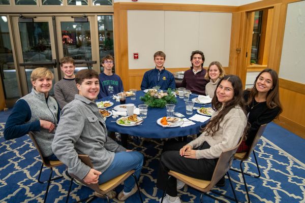 Group of young adults seated around a round table, smiling and enjoying a meal in a warmly lit, carpeted dining area.