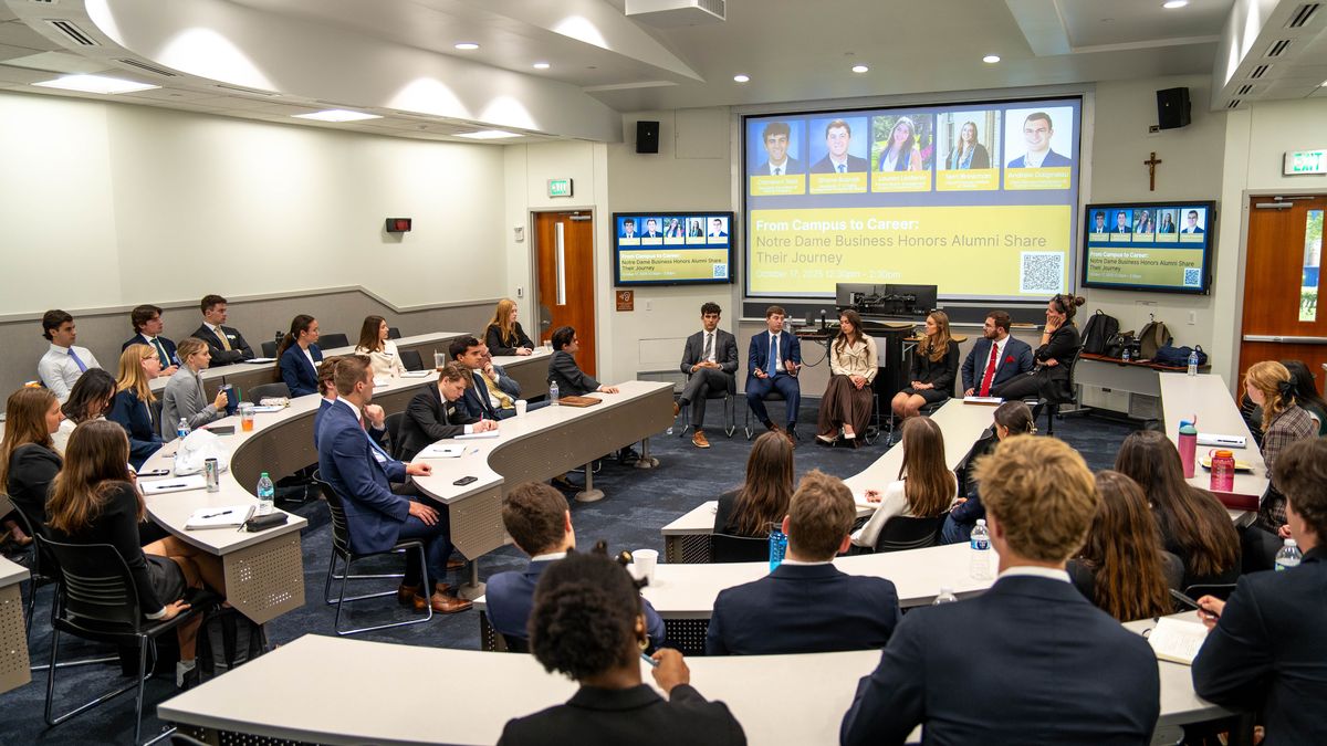 A panel of speakers is addressing a classroom filled with seated students. A screen displays information about a Notre Dame business alumni event.