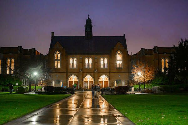 Illuminated Gothic-style building at night with arched windows, a wet pathway leading to it, and a purple sky in the background.