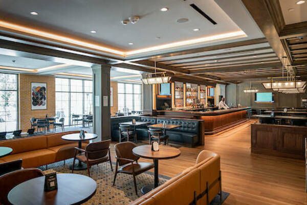 Modern restaurant interior with wooden floors, leather seating, and a central bar. Large windows provide natural light.