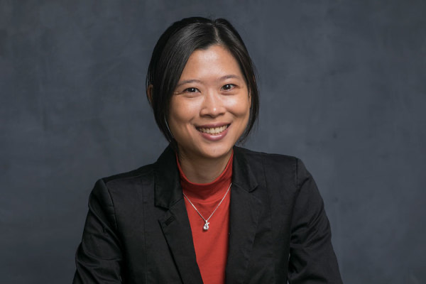 A woman with dark hair tied back, wearing a black jacket and red turtleneck, smiles against a dark gray background.