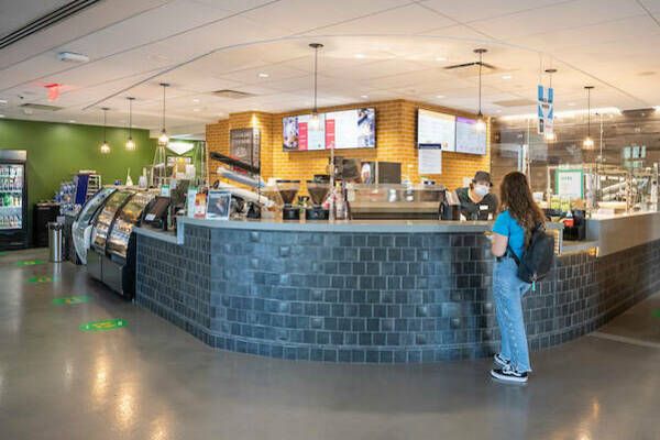 A student wearing a backpack speaks with a masked employee behind the counter at a modern campus café, which features digital menu boards, food displays, and a spacious interior.