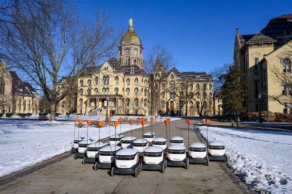 A row of delivery robots with orange flags lined up on a snowy path in front of a historic building with a golden dome under a clear blue sky.