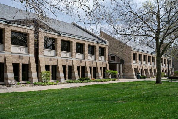 A brick academic building with large windows, surrounded by green grass and trees under a cloudy sky.