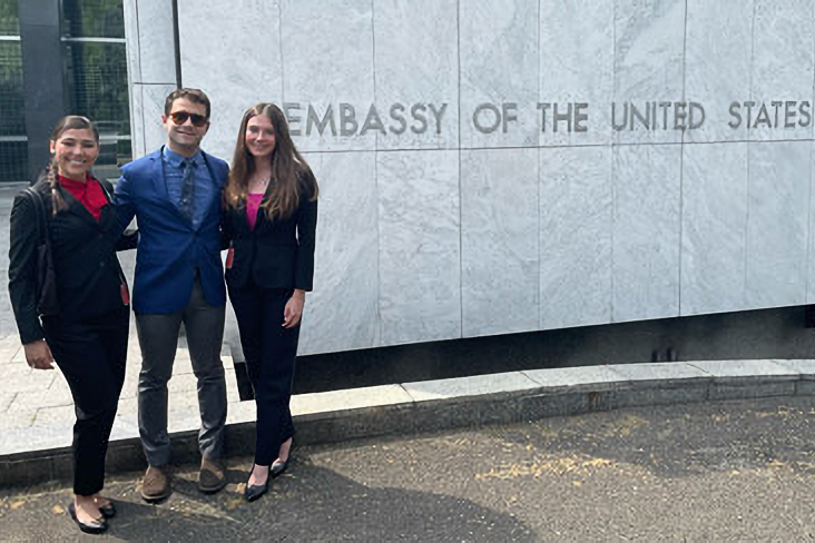 Three people standing in front of a U.S. Embassy building, smiling and dressed in business attire.
