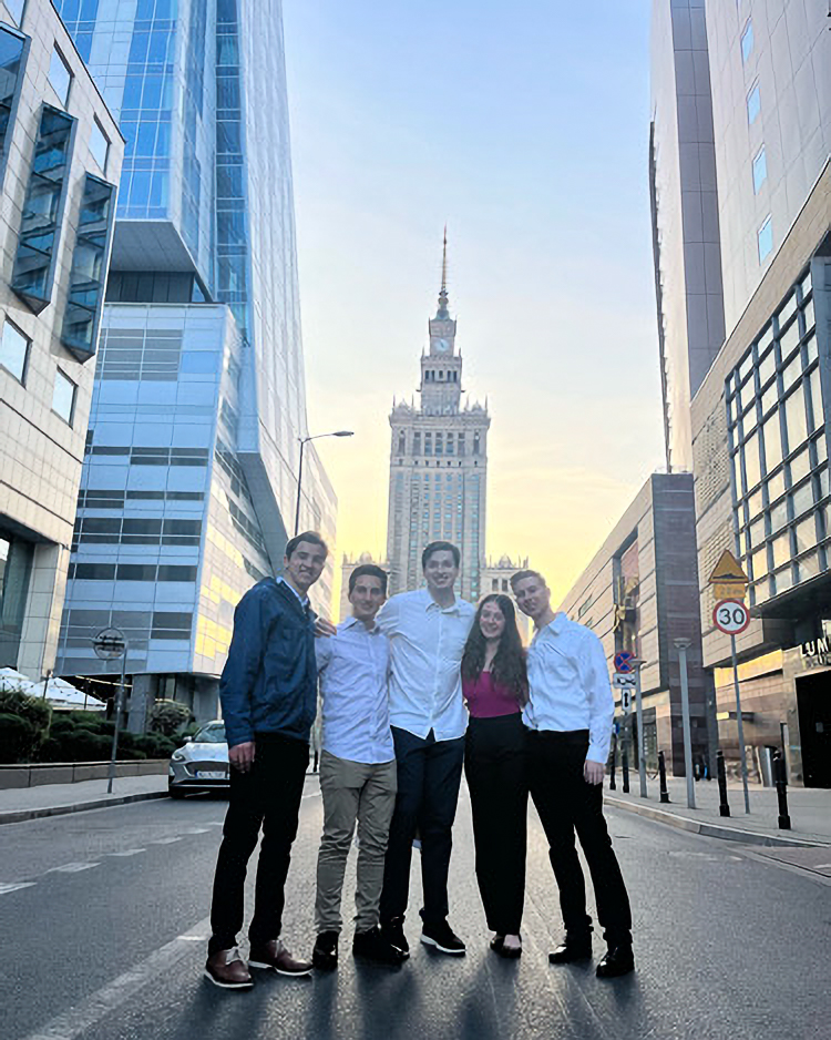 Five people stand smiling on a city street between tall buildings, with a tower in the background under a clear sky.