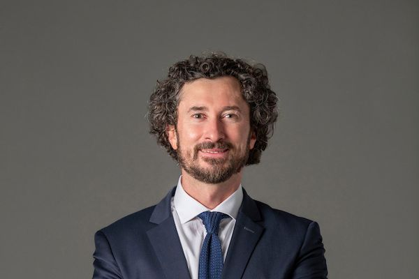 Man with curly hair in a suit and tie, smiling with arms crossed against a plain gray background.