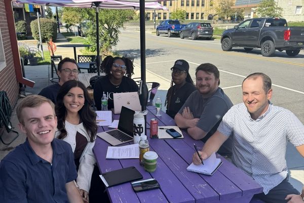 Group of people sitting around a purple picnic table outdoors, smiling, with laptops and drinks under a striped umbrella.