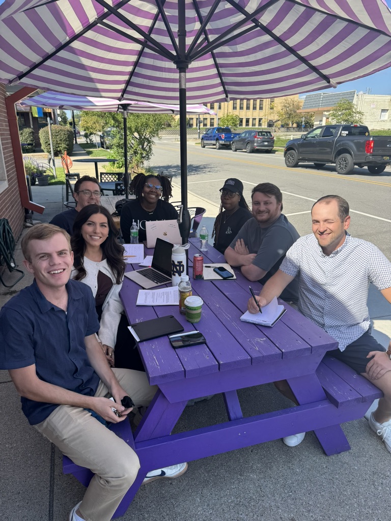 Team with Jas at co-op Group of people sitting around a purple picnic table with laptops and notepads, under a striped umbrella, in an outdoor setting.