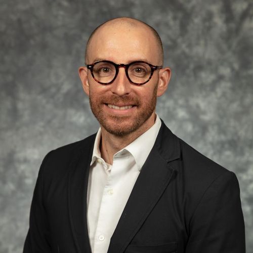 Jason Reed Headshot in front of gray and white fuzzy background wearing a black suit jacket with a white button up and he has glasses on